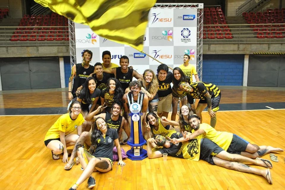 Estudantes da Atlética das Engenharias da UFU, vestidos de preto e amarelo, comemoram o título de 2019 ao redor do troféu.